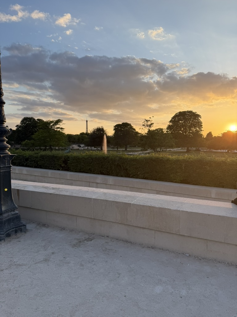 A scenic view of a Parisian park at sunset, featuring lush green trees, a fountain, and a distant view of the Eiffel Tower under a colorful sky.